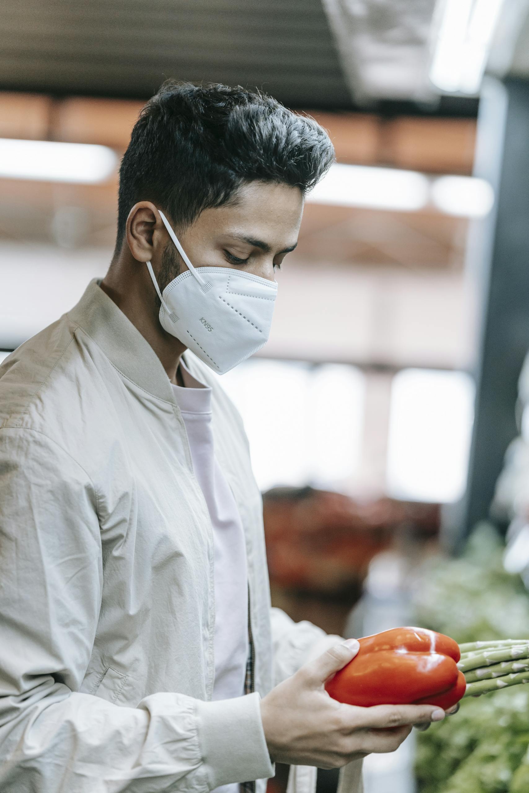 Side view of concentrated Indian male customer in medical mask standing with red bell pepper and asparagus in hands during grocery shopping on blurred background